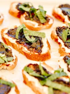 Plate full of slices of baguette with soft goat cheese spread on top, fig jam and mint leaf slices.