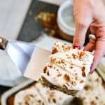 Hand serving a slice of pecan cake with the rest of the cake in the background.